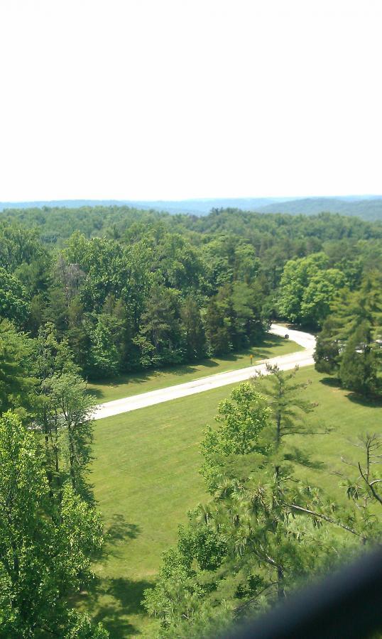 A scenic view of a winding road surrounded by lush green trees and open grassy areas, captured from above on a bright sunny day. The horizon features rolling hills in the background, creating a peaceful natural landscape. O'bannon Woods mountain bike trail.