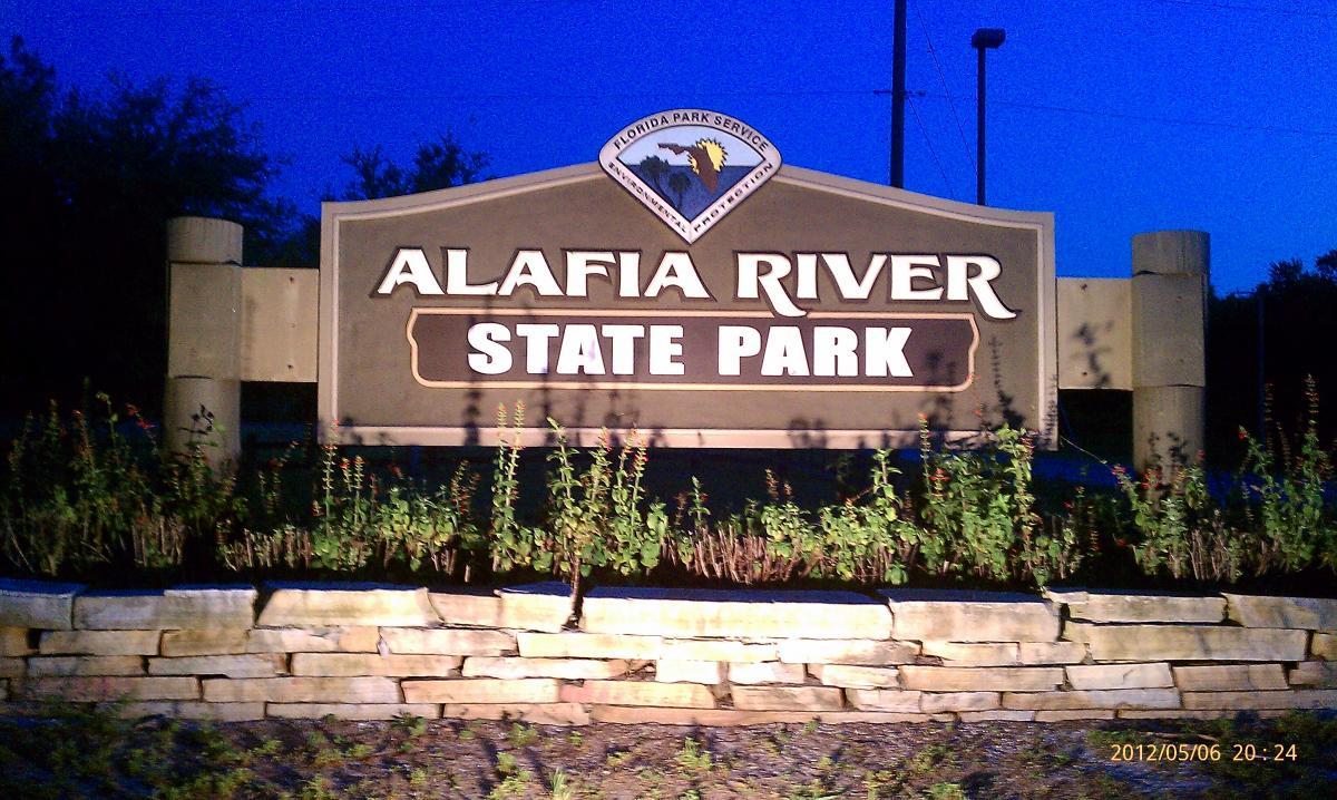 Sign for Alafia River State Park at dusk, featuring the park's name prominently displayed along with the Florida Park Service emblem, surrounded by a landscaped stone base and greenery. Alafia River State Park mountain bike trail.