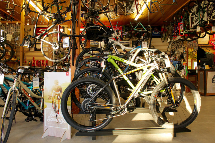 A busy bike shop interior featuring various bicycles on display. Several bikes are hung from the ceiling, while others are arranged on a stand in the foreground. A promotional poster for spring savings is prominently displayed, showcasing a cyclist. The shop is brightly lit, creating a welcoming atmosphere for customers.
