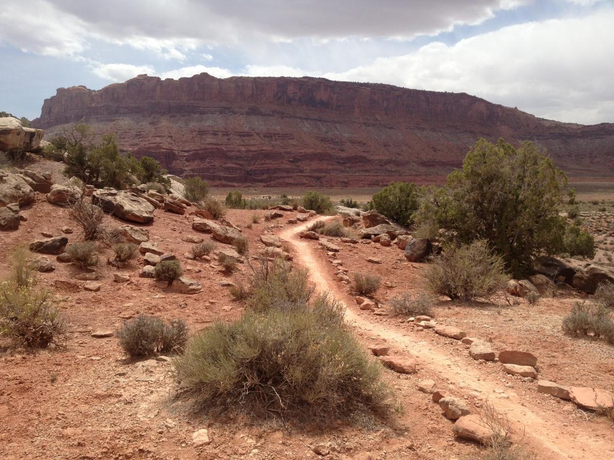 A winding dirt path leads through red rocky terrain, surrounded by sparse vegetation and shrubs, with a prominent layered rock formation in the background under a partly cloudy sky. Moab Brand Trails mountain bike trail.