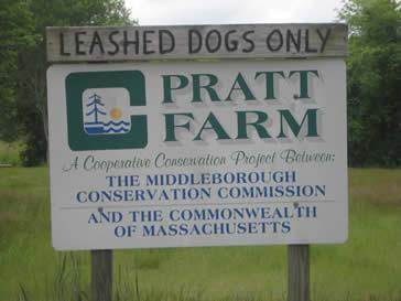 A sign for Pratt Farm, indicating that only leashed dogs are allowed. The sign also mentions that it is a cooperative conservation project between the Middleborough Conservation Commission and the Commonwealth of Massachusetts. Surrounding greenery is visible in the background. Pratt Farm mountain bike trail.