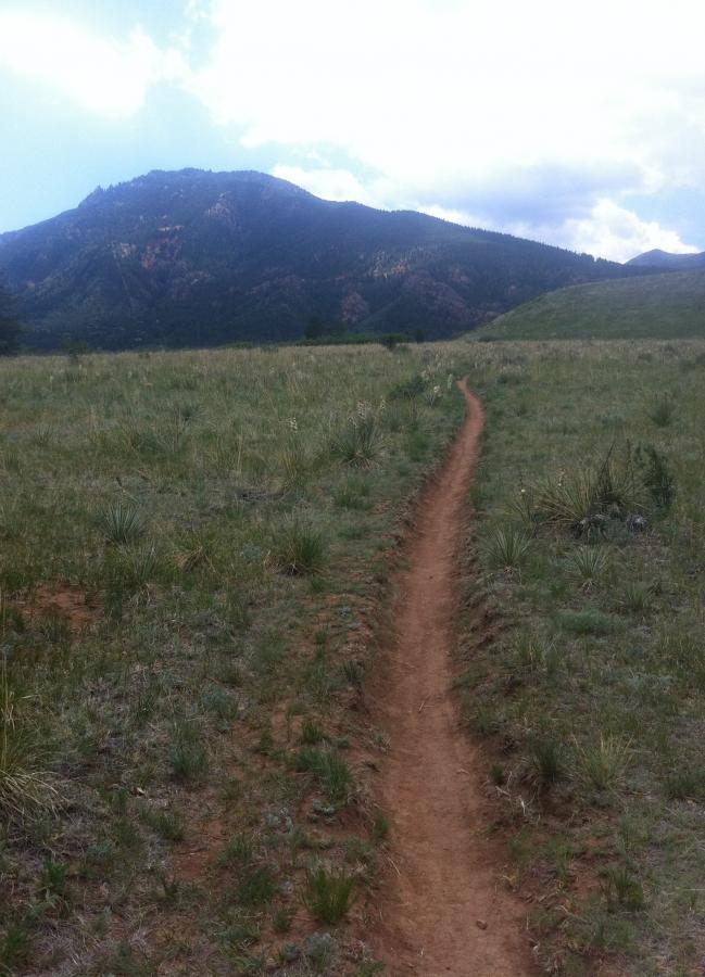A winding dirt trail leads through a grassy landscape, with a mountain rising in the background under a partly cloudy sky. The scene captures the natural beauty and serenity of a hiking path in a rural area. Stratton Open Space / The Chutes mountain bike trail.