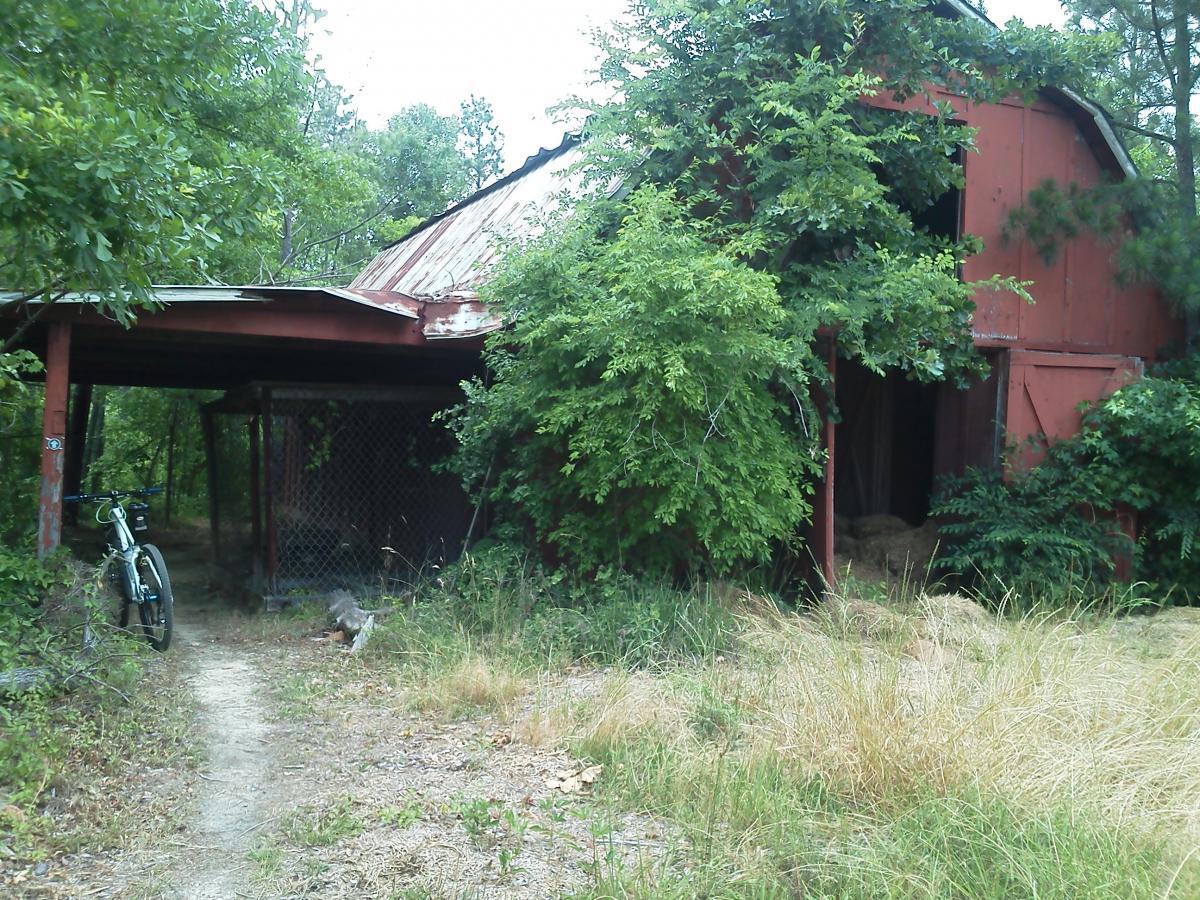 An old, weathered red barn partially covered in greenery, with a bicycle leaning against a post nearby. A dirt path leads toward the barn, surrounded by dry grass and trees. The roof shows signs of wear, indicating aging, while the entrance is partially open, revealing some hay inside. Arrowhead Park mountain bike trail.