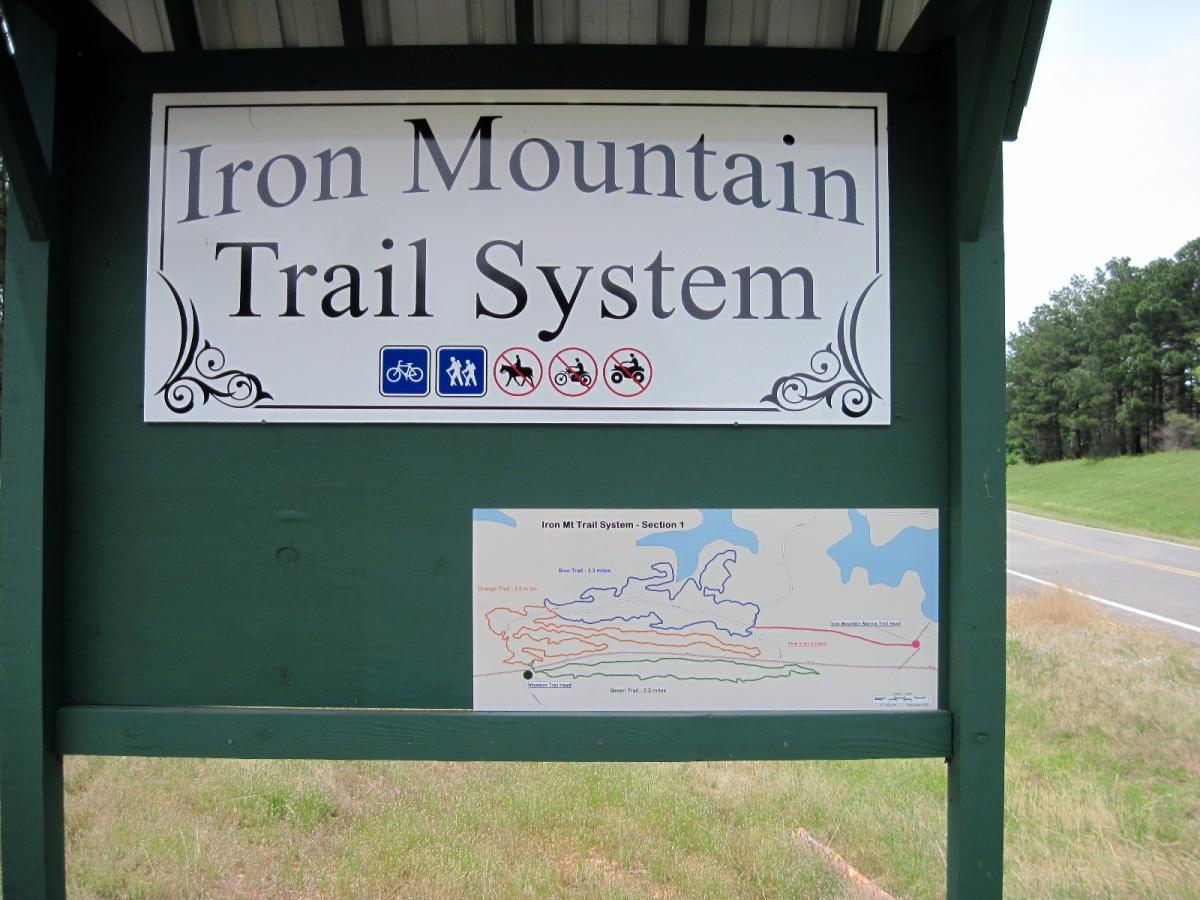 Sign for the Iron Mountain Trail System, featuring a map indicating different trails. Symbols for biking, hiking, and horseback riding are displayed above the sign, along with a prohibition symbol for motorized vehicles. The sign is positioned near a road, with trees in the background. Iron Mountain mountain bike trail.