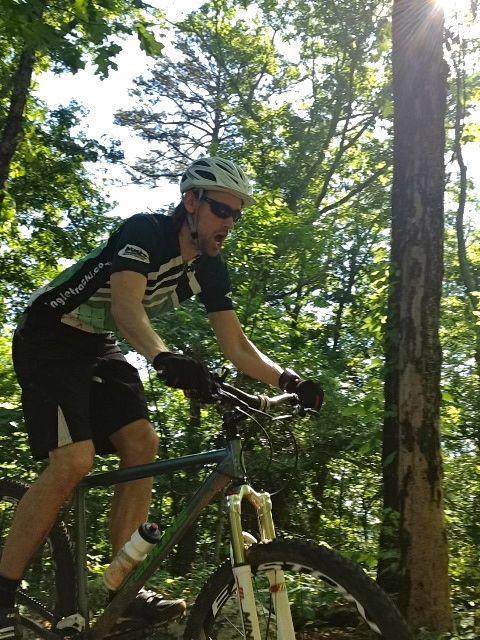 A cyclist riding a mountain bike on a sunny forest trail, surrounded by lush green trees. The cyclist is wearing a helmet and sunglasses, focused on navigating the terrain. Sunlight filters through the leaves, creating a vibrant and dynamic atmosphere. White Oak Mtn Biology Trails mountain bike trail.