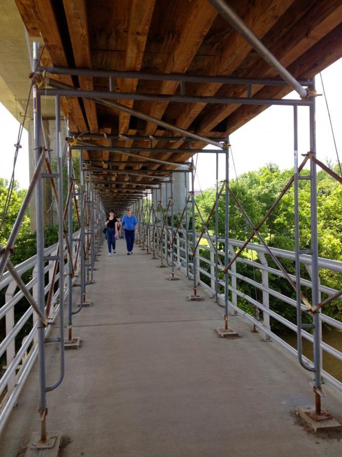 Two people walk along a walkway supported by scaffolding and wooden beams, with greenery visible alongside the path. The structure is part of an overpass or bridge, highlighting a mix of nature and construction elements. Belle Isle mountain bike trail.