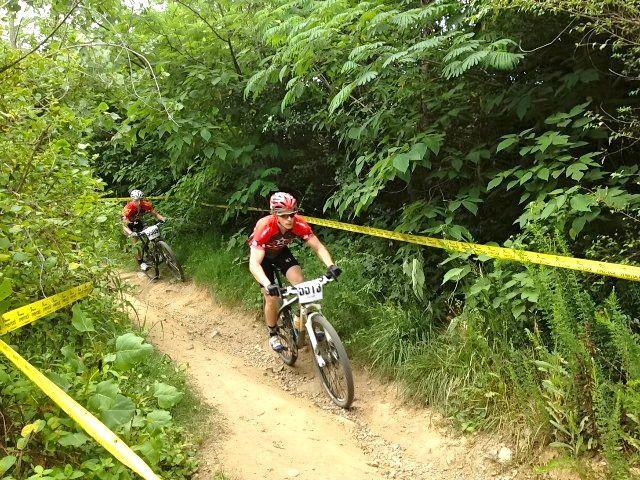 Two mountain bikers navigate a narrow dirt trail surrounded by dense greenery. The riders, both wearing red jerseys, are focused on the path ahead as they maneuver around yellow caution tape marking the course. Northbank Trail mountain bike trail.