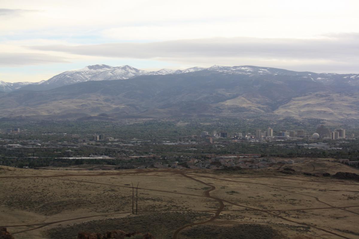 A panoramic view of a city surrounded by mountains, showcasing a mixture of urban and natural landscapes. The foreground features a dry, grassy terrain with dirt paths, while the background displays snow-capped peaks under a cloudy sky. The city skyline includes several tall buildings and structures nestled among green trees. Chipset Trail mountain bike trail.