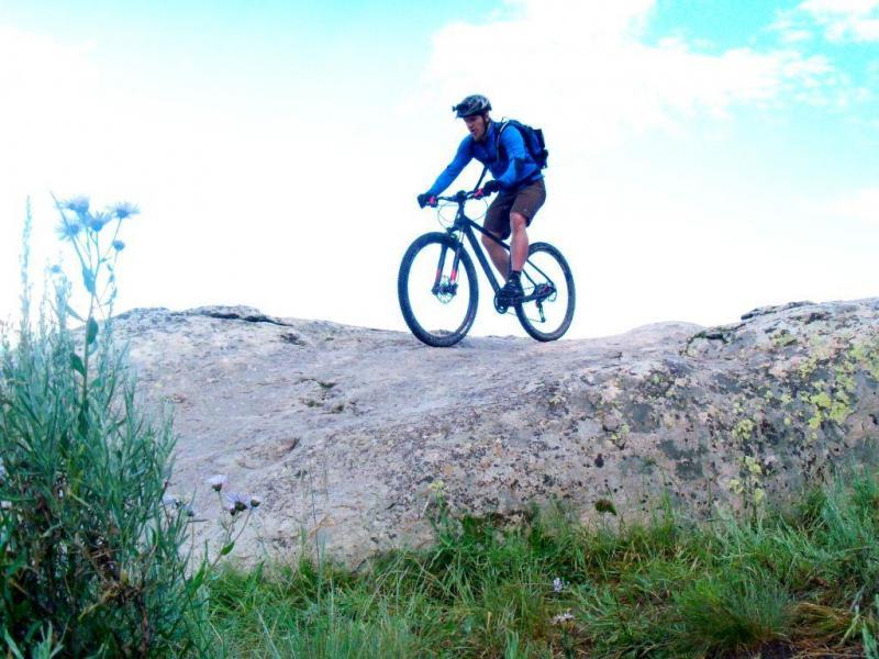Ibex Maroc 29: A mountain biker navigating over large rocks, wearing a helmet and a blue shirt, with a backpack, surrounded by greenery and a clear sky.