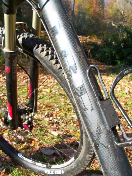 Ibex Maroc 29: Close-up view of a mountain bike's carbon fiber frame and front fork, with a water bottle cage attached. The ground is covered in fallen leaves, indicating an outdoor environment. The bike features rugged tires suitable for off-road terrain.