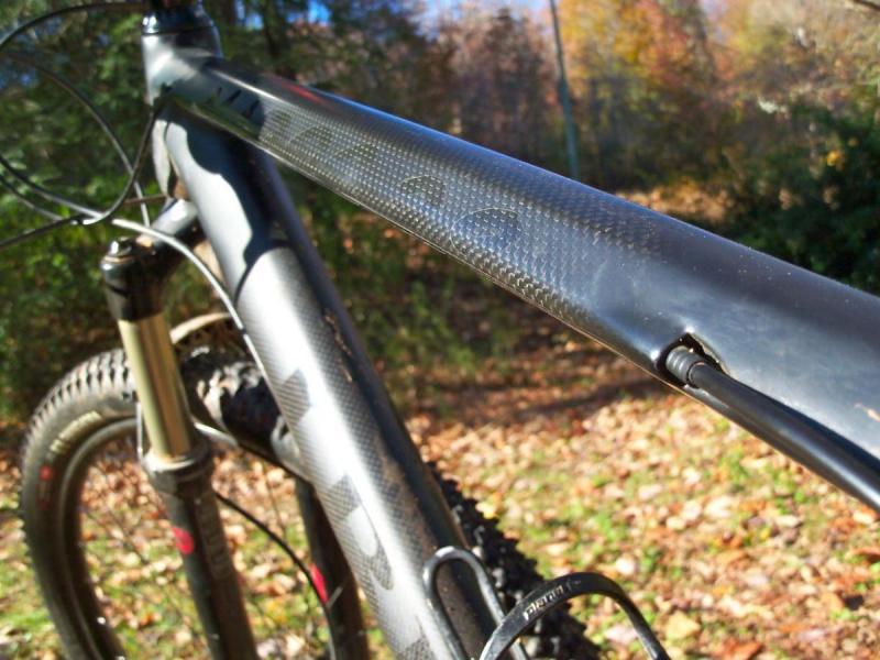 Ibex Maroc 29: Close-up view of a mountain bike's frame, showcasing a sleek carbon fiber top tube and part of the front suspension. The background features a forested area with autumn foliage on the ground.