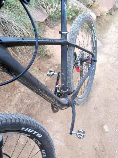 Ibex Maroc 29: A close-up view of a mountain bike's rear frame and drivetrain, showing the pedals, chain, and tire on a dirt trail with some vegetation in the background.