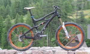 Ibis Mojo HD: A mountain bike with a black frame and orange wheels is positioned on a stone ledge, set against a backdrop of green trees and a hilly landscape. The bike features a suspension fork and a racing saddle, emphasizing its design for off-road riding.