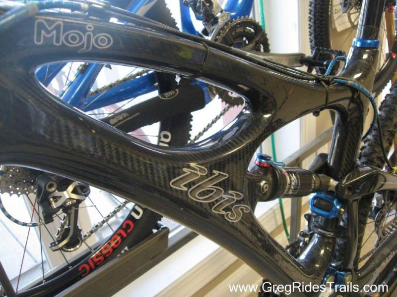 Ibis Mojo HD: Close-up view of a black Ibis Mojo mountain bike frame showcasing its carbon fiber construction and design details, including the brand name and suspension components. The background features additional bicycles.