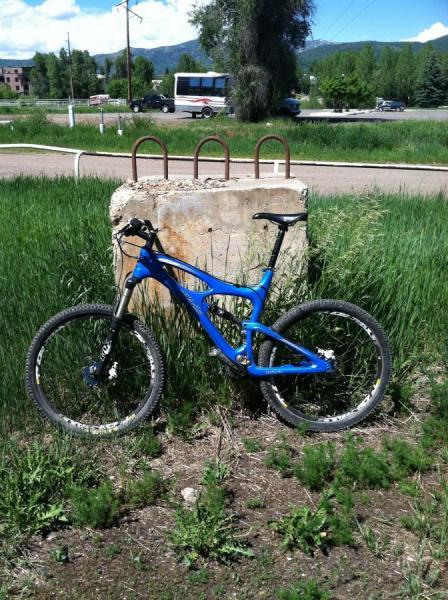 Ibis Mojo SL: A blue mountain bike is parked next to a large concrete block surrounded by tall green grass, with a road and trees visible in the background.