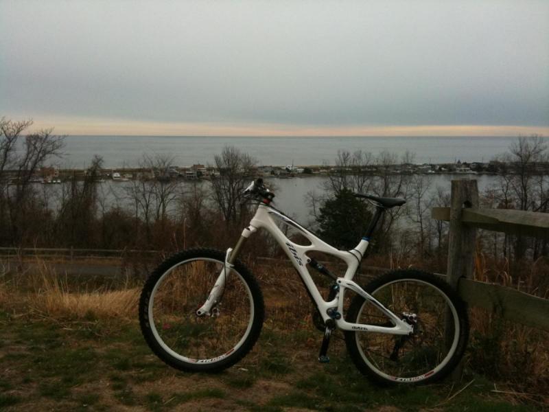 Ibis Mojo SL: A mountain bike parked on a grassy hillside, overlooking a calm lake with a cloudy sky in the background. The shoreline features a small marina with boats, surrounded by bare trees.