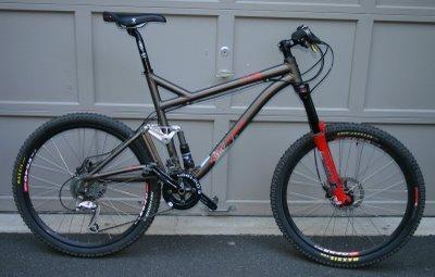 A modern mountain bike featuring a sleek aluminum frame with a combination of black and metallic brown colors. The bike is equipped with front and rear suspension, wide tires, disc brakes, and a prominent crankset. It is positioned next to a garage door, showcasing its design and features.