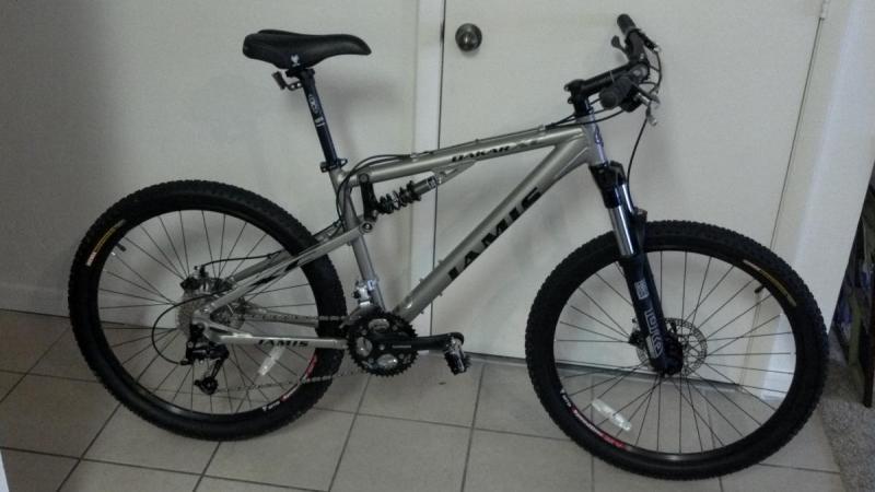 Jamis Dakar XC: A silver mountain bike with thick tires, front suspension, and a black seat, positioned against a wall with a closed door in the background. The bike features a simple, modern design and appears to be in good condition.