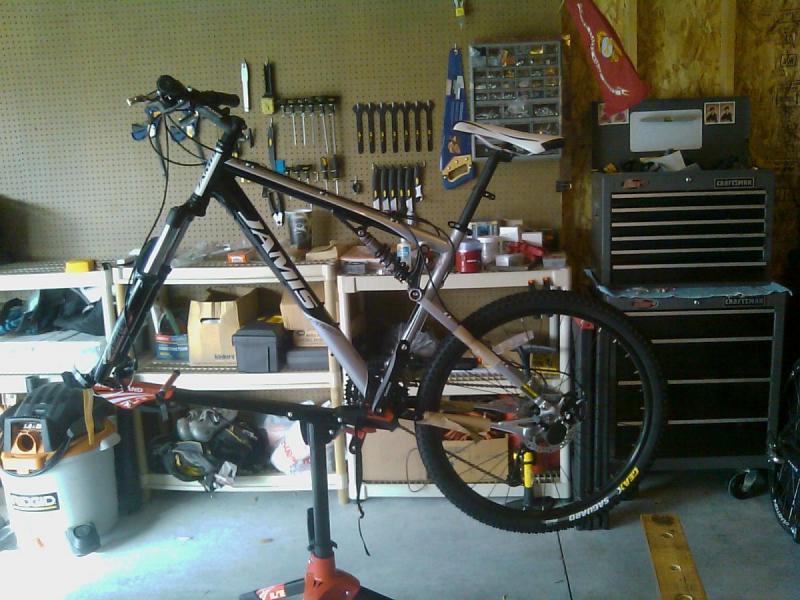 Jamis Dakar XC: A mountain bike is mounted on a repair stand in a garage workshop. The bike features a black and white frame with visible brand markings. Behind it, there are shelves lined with various tools and equipment, including wrenches, screwdrivers, and other cycling accessories. A toolbox and a portable device are visible on the floor, along with other cycling gear and supplies. The setting is cluttered but organized, reflecting a space dedicated to bike maintenance and repairs.