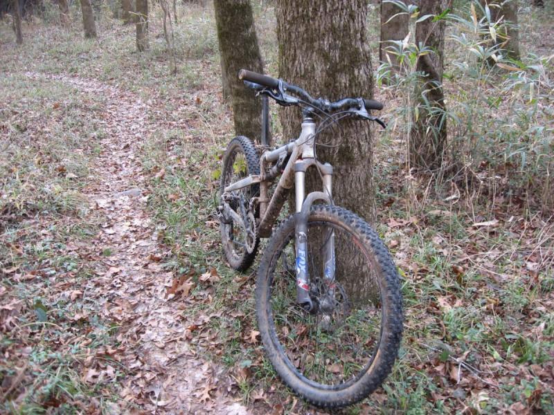 Jamis Dakar XLT: A mountain bike leaning against a tree on a forest trail, surrounded by fallen leaves and grass. The bike appears to be used and muddy, indicating it has been on a rugged outdoor ride.