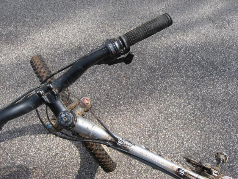 Jamis Dakar XLT: Top view of a mountain bike's handlebars, showing the grips, brake levers, and gear shifters, with dirt and wear visible on the components. The bike is positioned on an asphalt surface.