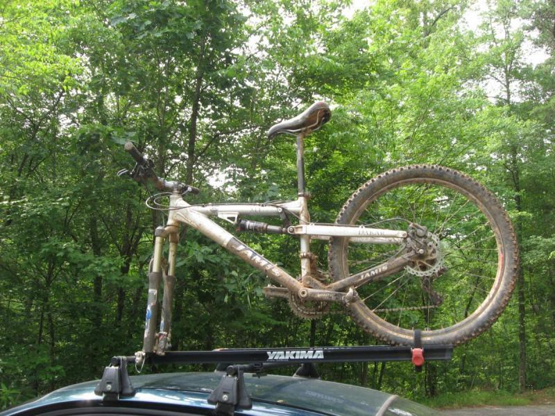 Jamis Dakar XLT: A mountain bike mounted on the roof rack of a car, surrounded by lush green trees. The bike appears muddy, indicating recent use on off-road trails. The roof rack is labeled with the brand name "Yakima."