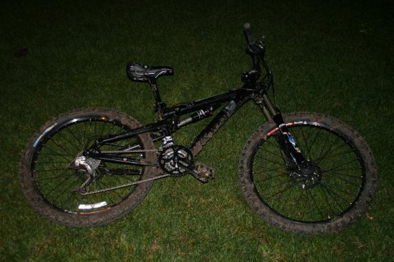 Jamis Dakar XLT: A black mountain bike with muddy tires is resting on green grass. The bike features a sturdy frame, front suspension forks, and a single gear setup. The scene is set at night, with limited visibility.