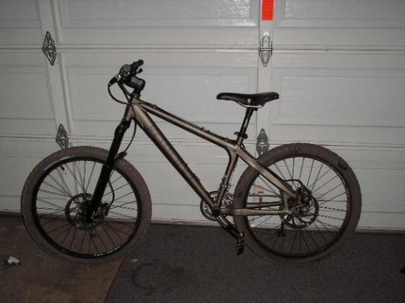 Jamis Komodo 2.0: A mountain bike with a gray frame, front suspension, and disc brakes, positioned against a garage door. The bike features thick tires suitable for off-road terrain and has a simple, streamlined design.