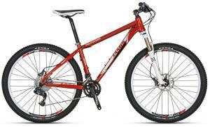 Jamis Nemesis: A red mountain bike with a black seat and handlebars, featuring front suspension and disc brakes. The bike has multiple gears and is designed for off-road cycling.