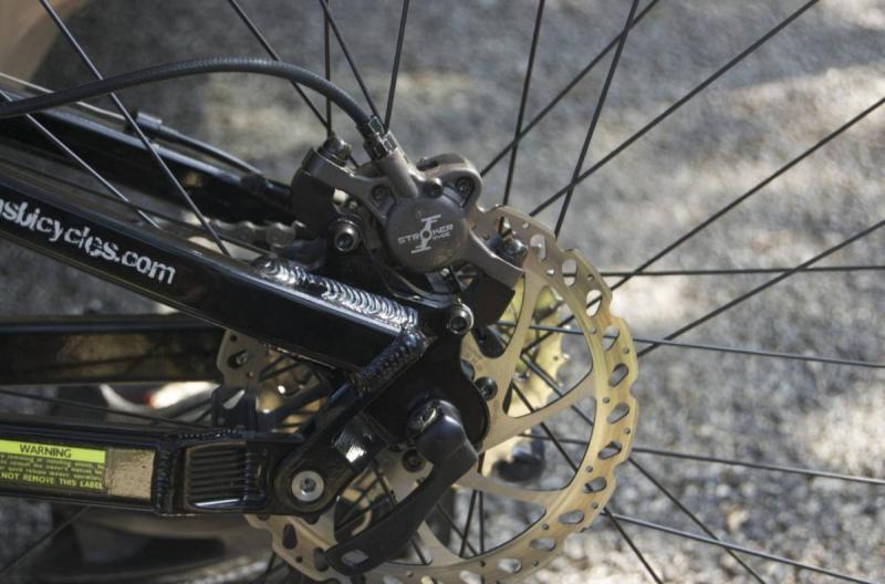 KHS XCT 555: Close-up view of a bicycle’s rear wheel, focusing on the disc brake system. The image shows the brake caliper, rotor, and part of the bike frame, highlighting the intricate details of the braking mechanism against a blurred gravel background.