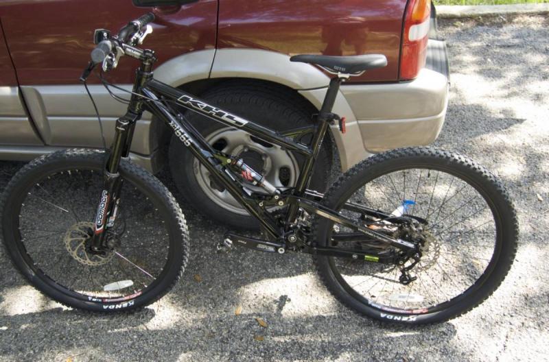 KHS XCT 555: A black mountain bike parked beside a car on a gravel surface. The bike features a suspension system and knobby tires, with visible branding on the frame and wheels. The car in the background is a dark red color, partially obscured by the bike.