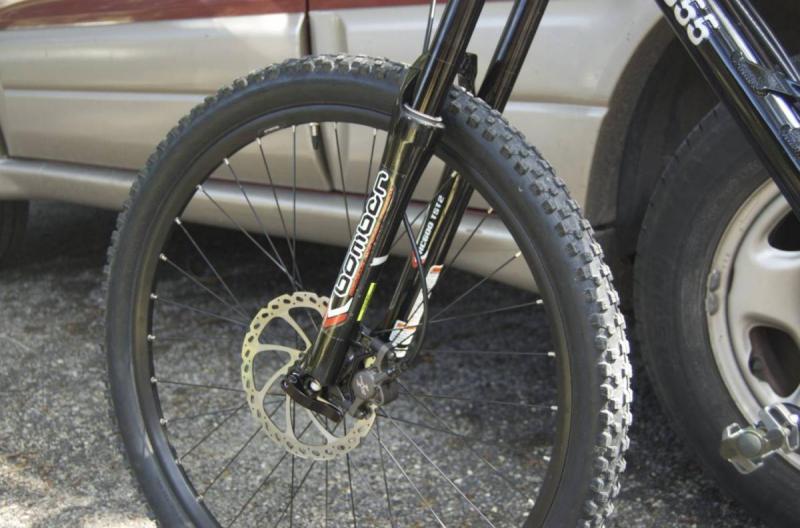 Kenda Nevegal: Close-up view of a mountain bike's front wheel and fork, featuring a black frame and a disc brake. In the background, a silver car is partially visible, parked on a gravel surface.