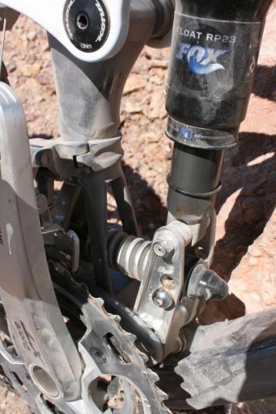 Kona Abra Cadabra: Close-up view of a mountain bike's rear suspension system, showcasing the shock absorber, linkage, and chainring. The components are dusty, indicating use in an outdoor setting.