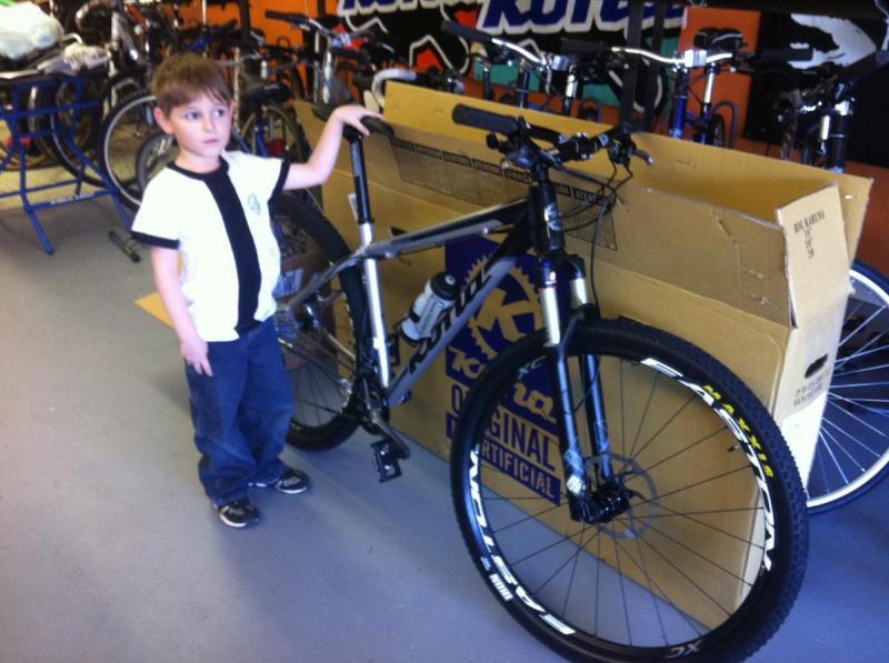Kona Big Kahuna: A young boy stands next to a mountain bike in a bicycle shop, with a large cardboard box in the background. The boy has short brown hair and is wearing a white shirt with a black stripe and blue jeans. The bike is silver with black wheels and a water bottle mounted on the frame. Various bicycles are visible in the shop behind him.