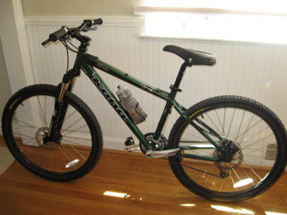Kona Blast: A green mountain bike with thick tires, a bottle holder, and front suspension, positioned on a wooden floor in a well-lit room. The bike features a minimalist design and is leaning against a white wall with natural light coming through a nearby window.