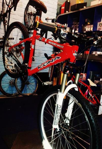 Kona Cinder Cone: A red Kona mountain bike is positioned prominently in a bike shop, with another bike partially visible in the background. The bike features distinctive handlebars, a suspension fork, and various components. The shop interior includes shelves with bike accessories and equipment.