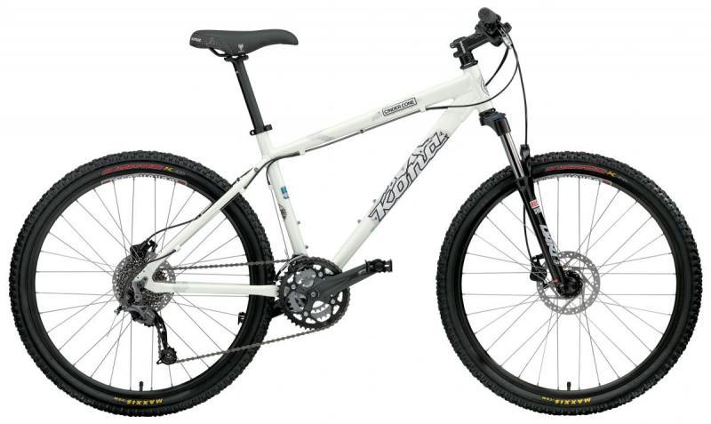 Kona Cinder Cone: A white mountain bike with a sleek design, featuring wide black tires, a front suspension fork, and multiple gears. It has a straight handlebar, disc brakes, and is positioned upright on a plain background.