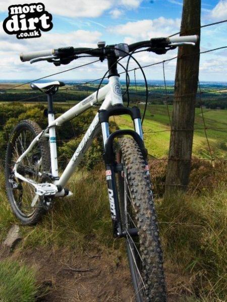 Kona Cinder Cone: Close-up view of a mountain bike resting near a fence on a grassy hillside. The bike is white with black and silver components, showcasing its front fork and handlebar. In the background, a scenic landscape features rolling hills under a partly cloudy sky.