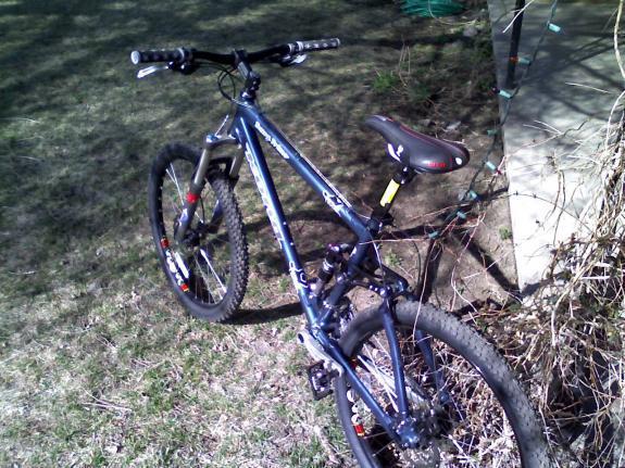 Kona Dawg Primo: A blue mountain bike resting on the grass beside a building, partially hidden by shrubs. The bike features thick tires, a sturdy frame, and detailed handlebars. Sunlight is casting shadows on the ground, indicating a clear day.