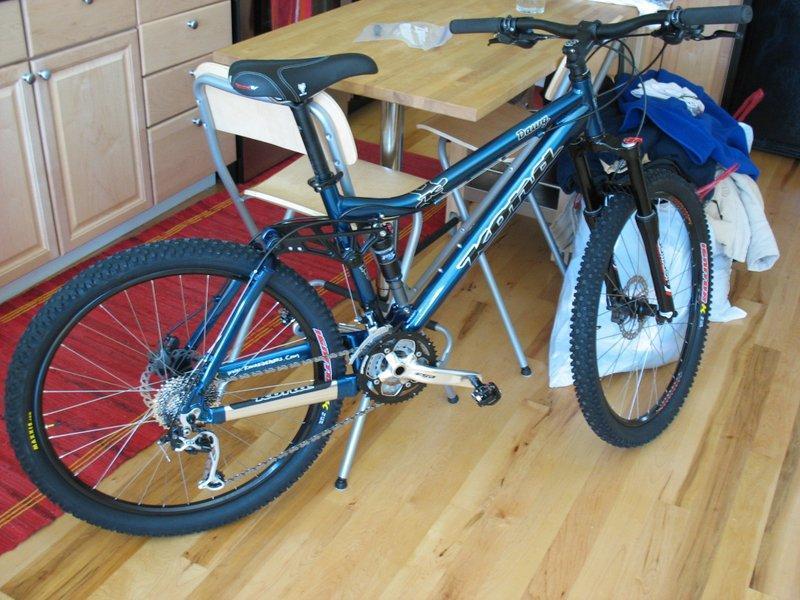 Kona Dawg: A blue mountain bike with thick tires is positioned next to a wooden dining table and chairs in a room with light wood flooring. Behind the bike, there are items draped over a chair, including a blue shirt and other clothing. The background features a kitchen with wooden cabinets, and a red area rug is visible on the floor.