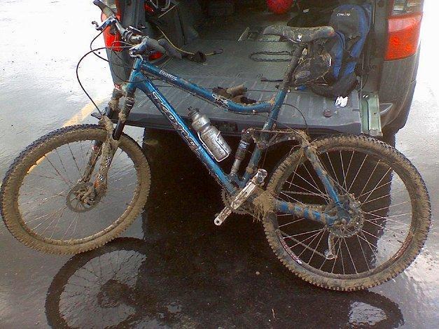 Kona Dawg: A muddy mountain bike leaning against the open back of a vehicle, with dirt visible on the frame, tires, and parts. A water bottle is attached to the frame, and the background shows the interior of the vehicle and a wet parking surface.