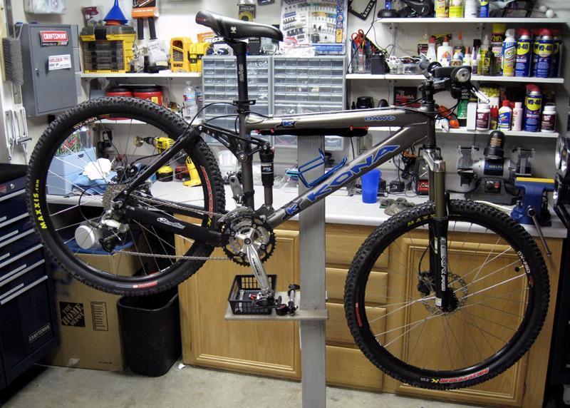 Kona Dawg: A Kona mountain bike is displayed on a repair stand in a workshop, surrounded by various tools and equipment. The bike features black and blue details with Maxxis tires, and the background includes organized shelves filled with tools and maintenance supplies.