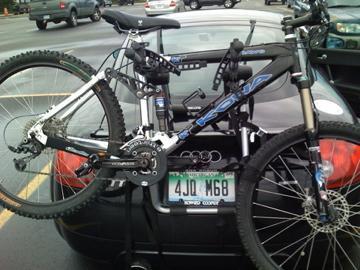 Kona Dawg: A mountain bike securely mounted on the rear of a black car in a parking lot. The bike features a silver and blue frame, with visible gears and tires. The car has a visible license plate and is surrounded by other vehicles in the background.
