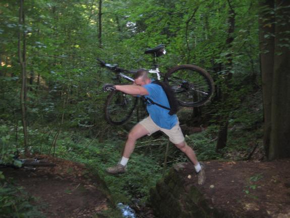 Kona Fire Mountain Deluxe: A person in a blue t-shirt and shorts leaps over a small gap in a forested area, carrying a mountain bike on their back. The surrounding greenery includes trees and shrubs, creating a natural, outdoor setting.