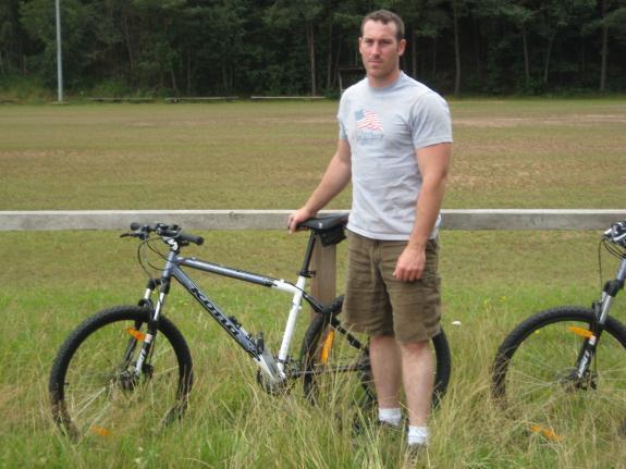Kona Fire Mountain Deluxe: A man standing next to a mountain bike in a grassy field, wearing a gray t-shirt with an American flag design and khaki shorts. There are two bikes visible, and trees line the background.
