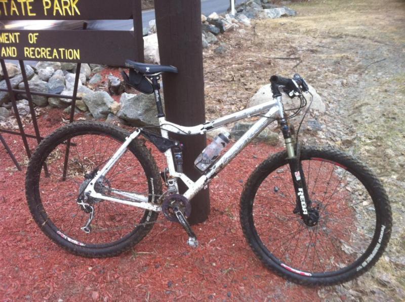 Kona Hei Hei 2-9: A mountain bike with a white frame, covered in dirt, parked next to a signpost in front of a state park. The bike has thick tires and a water bottle attached to the frame, with rocky terrain and mulch in the background.