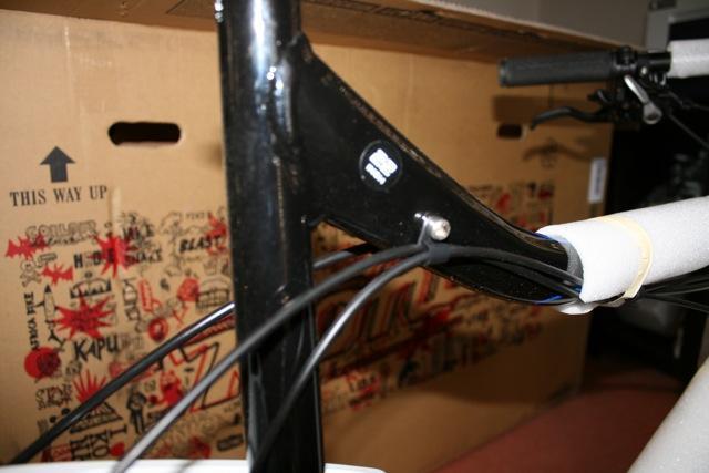 Kona Hei Hei 2-9: Image of a bicycle frame with a close-up view of the black metal tubing and handlebars. The frame features visible wires for brakes or shifting. In the background, a cardboard box with colorful graphics and the words "THIS WAY UP" is partially visible, suggesting the bicycle is newly unpacked.