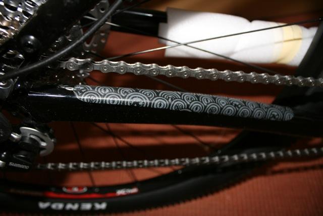 Kona Hei Hei 2-9: Close-up view of a bicycle's chainstay, featuring a patterned protective wrap with concentric circles, and showing parts of the drivetrain including the chain and rear cassette. The image highlights the bike's sleek black frame and components.