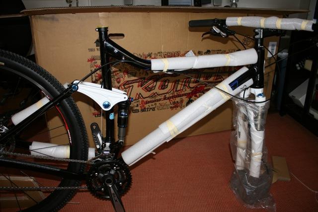 Kona Hei Hei 2-9: A partially assembled mountain bike with protective padding on its frame and components, positioned next to a large cardboard box that features brand logos and images. The bike is set on a carpeted floor.