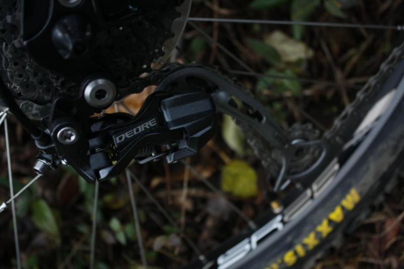 Kona Tanuki: Close-up image of a bicycle's rear derailleur, prominently featuring the Shimano Deore brand, set against a backdrop of foliage and leaves. The derailleur is shown in detail, highlighting its mechanical components and connection to the bike's chain.